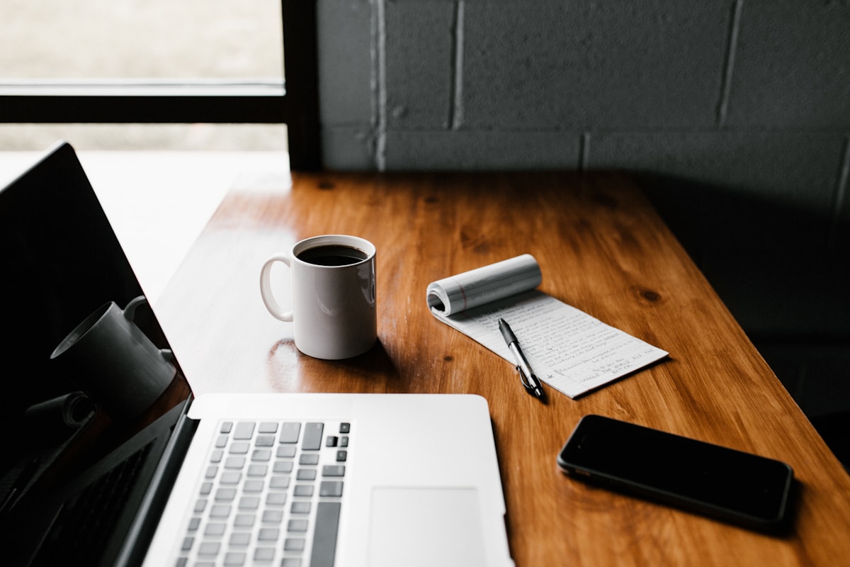 A laptop, phone, notebook, and coffee on a wooden desk